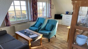 A room with a sofa, two armchairs, a coffee table, a TV, a white dresser, and a window with curtains. - Watt'n Blick, 70sqm for up to 4 people - Haus am Wattenmeer (Sylt)