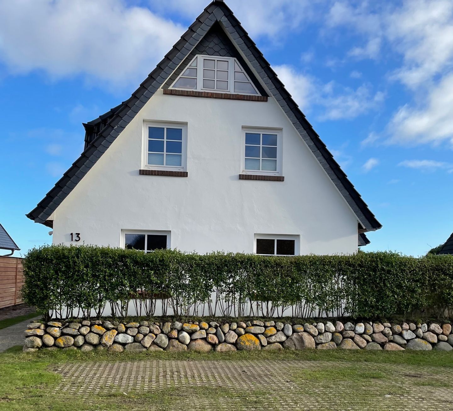 A white two-story house with a gabled roof, numbered 13, surrounded by a hedge and a stone wall.