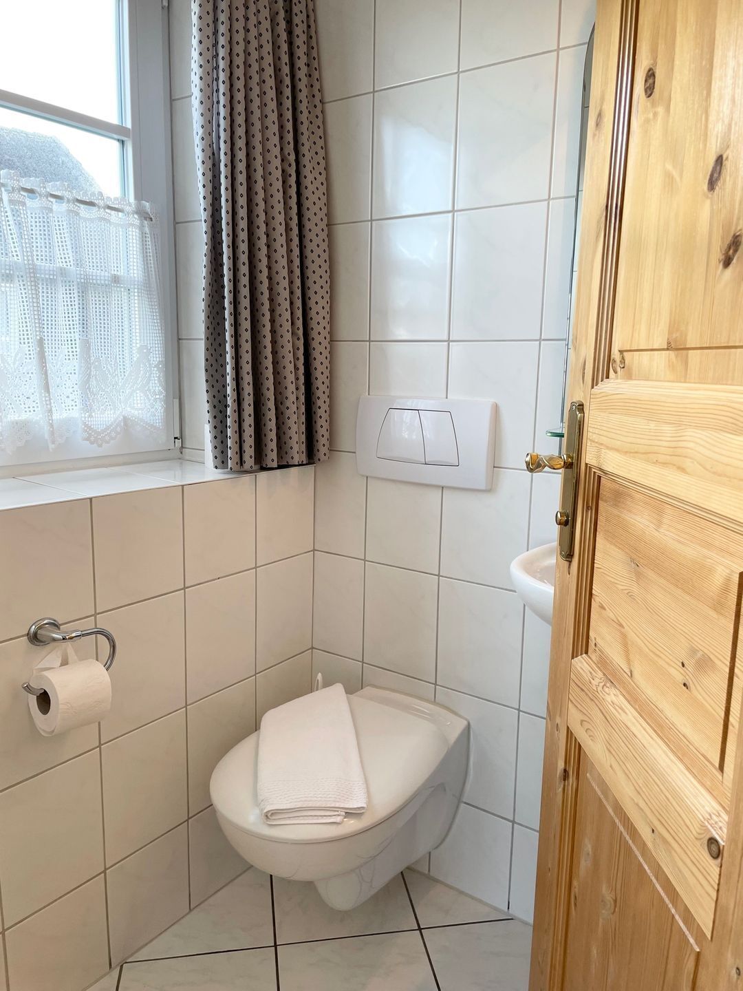 A bathroom with a toilet, a window with curtains, a wall-mounted light switch, and a wooden door.