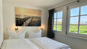 A hotel room with two beds, a painting of a sunset over a pier, a window with a view of greenery, and a curtain. - Wadden runner, 40sqm for up to 2 people - Haus am Wattenmeer (Sylt)