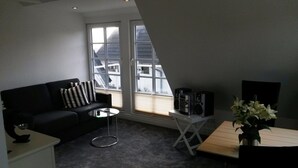 A modern living room with a black sofa, a glass coffee table, a small white table, and a plant. There are large windows with a view of buildings outside. - Wadden runner, 40sqm for up to 2 people - Haus am Wattenmeer (Sylt)