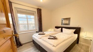 A bedroom with a large bed, a window with curtains, a radiator, and a bedside table with a lamp. - Watt'n Idyll, 50sqm for up to 2 people - Haus am Wattenmeer (Sylt)