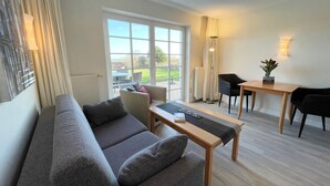 A modern living room with a grey sofa, a wooden coffee table, and a dining area with a table and chairs. - Watt'n Idyll, 50sqm for up to 2 people - Haus am Wattenmeer (Sylt)