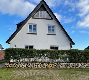 Een wit, twee verdiepingen tellend huis met een puntige dakkapel, nummer 13, omringd door een stenen muur en een haag.