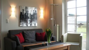 A living room with a grey sofa, red cushions, a wooden coffee table, and a painting on the wall. - Watt'n Idyll, 50sqm for up to 2 people - Haus am Wattenmeer (Sylt)