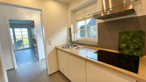 A modern kitchen with white cabinets, a stainless steel range hood, a black induction cooktop, and a window with blinds. - Watt'n Idyll, 50sqm for up to 2 people - Haus am Wattenmeer (Sylt)