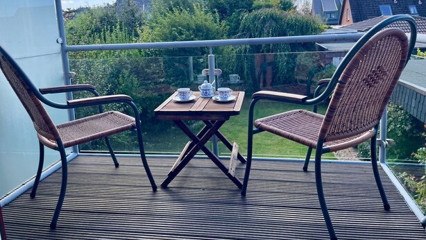 A balcony with two wicker chairs and a small table, overlooking a garden and buildings.