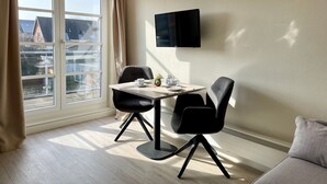 A modern room with a dining table and chairs, a flat-screen TV mounted on the wall, and a large window with curtains. - Starfish, 30sqm for up to 2 people - Haus an den Wiesen (Sylt-Ost)