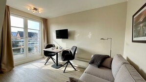 A modern living room with a sofa, two chairs, a dining table, a flat-screen TV, and a large window with curtains. - Starfish, 30sqm for up to 2 people - Haus an den Wiesen (Sylt-Ost)