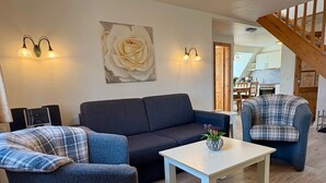 Un salon avec un canapé foncé, un fauteuil à motifs écossais, une table basse blanche et un tableau représentant une rose accroché au mur.