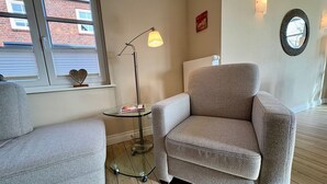 Ein Zimmer mit zwei Sesseln, einem kleinen Glastisch, einer Stehlampe und einem Fenster mit Jalousien.