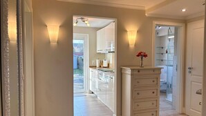 Ein Flur mit Holzboden, einem weißen Kommode mit Schubladen und einer Vase mit Blumen. Wandleuchten sorgen für Licht.
