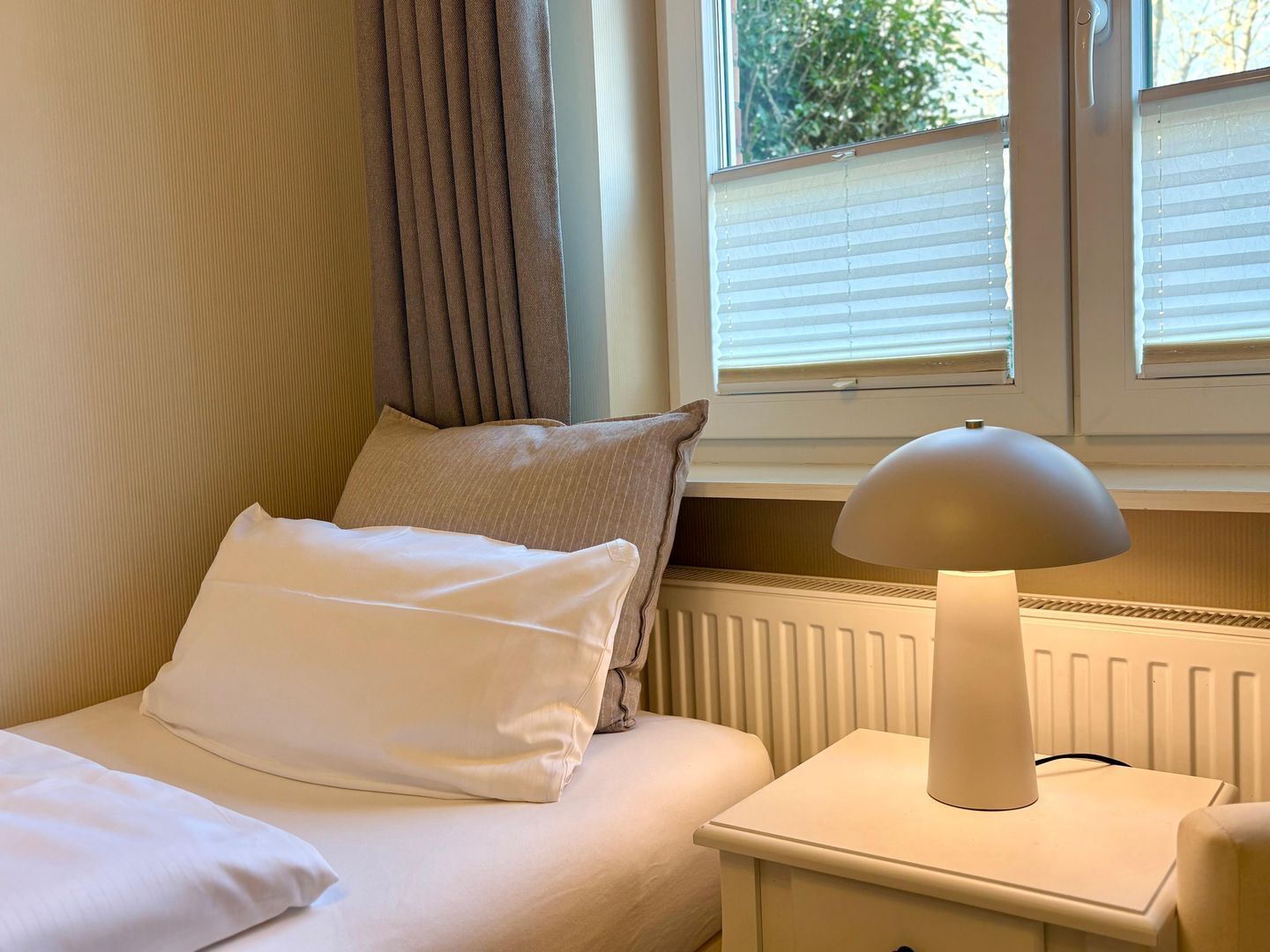 Une chambre à coucher avec un lit, des oreillers, une table de chevet, une lampe et une fenêtre avec des stores.