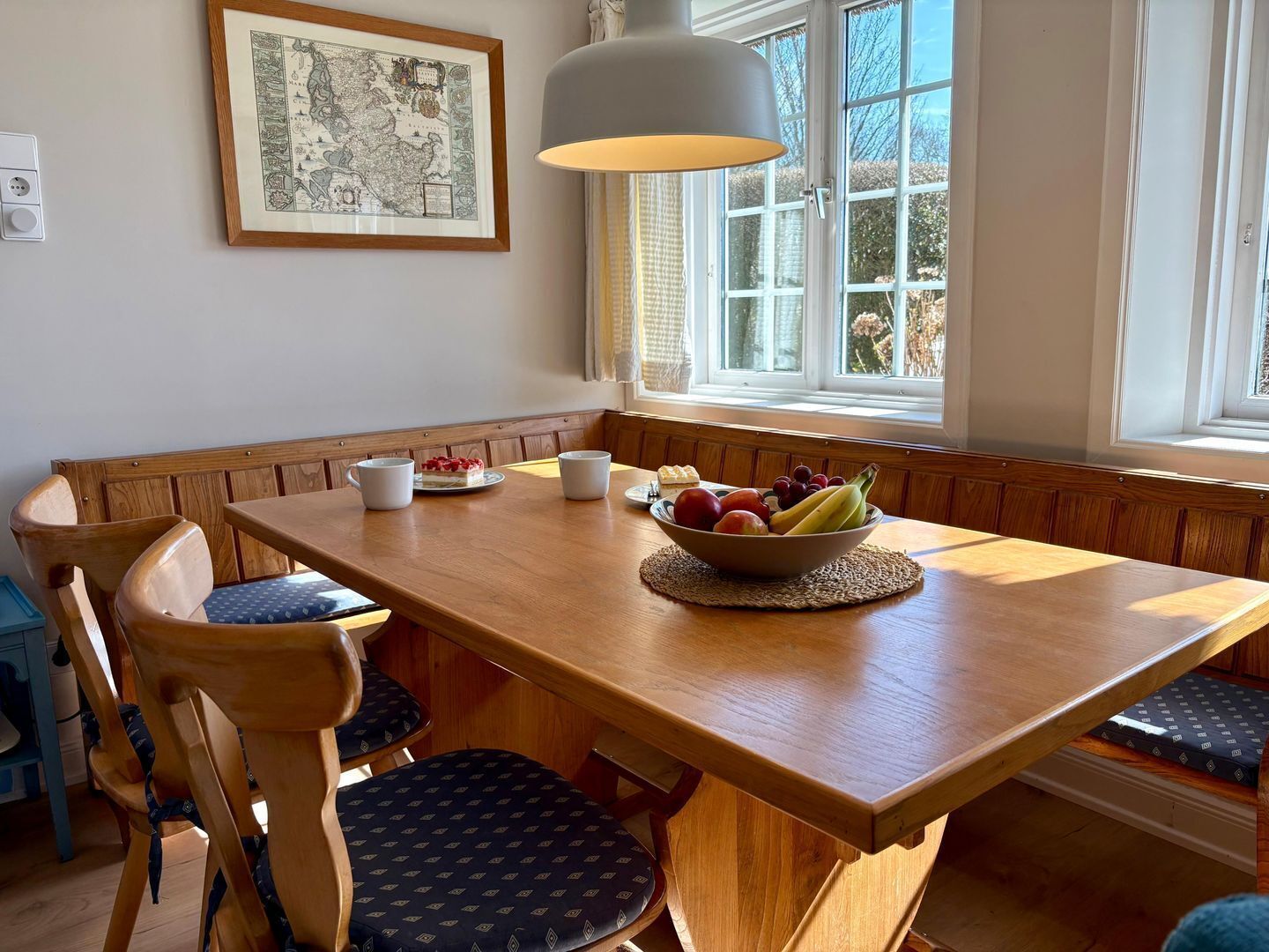 A dining area with a wooden table, bench seating, a bowl of fruit, and a framed map on the wall.