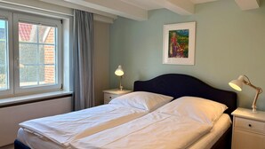 Une chambre à coucher comprenant un lit, deux tables de chevet, une fenêtre avec des rideaux, un tableau au mur et un plafond avec des poutres apparentes.