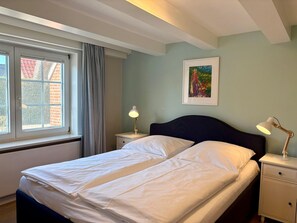 Une chambre à coucher comprenant un lit, deux tables de chevet, une fenêtre avec des rideaux, un tableau au mur et un plafond avec des poutres apparentes.