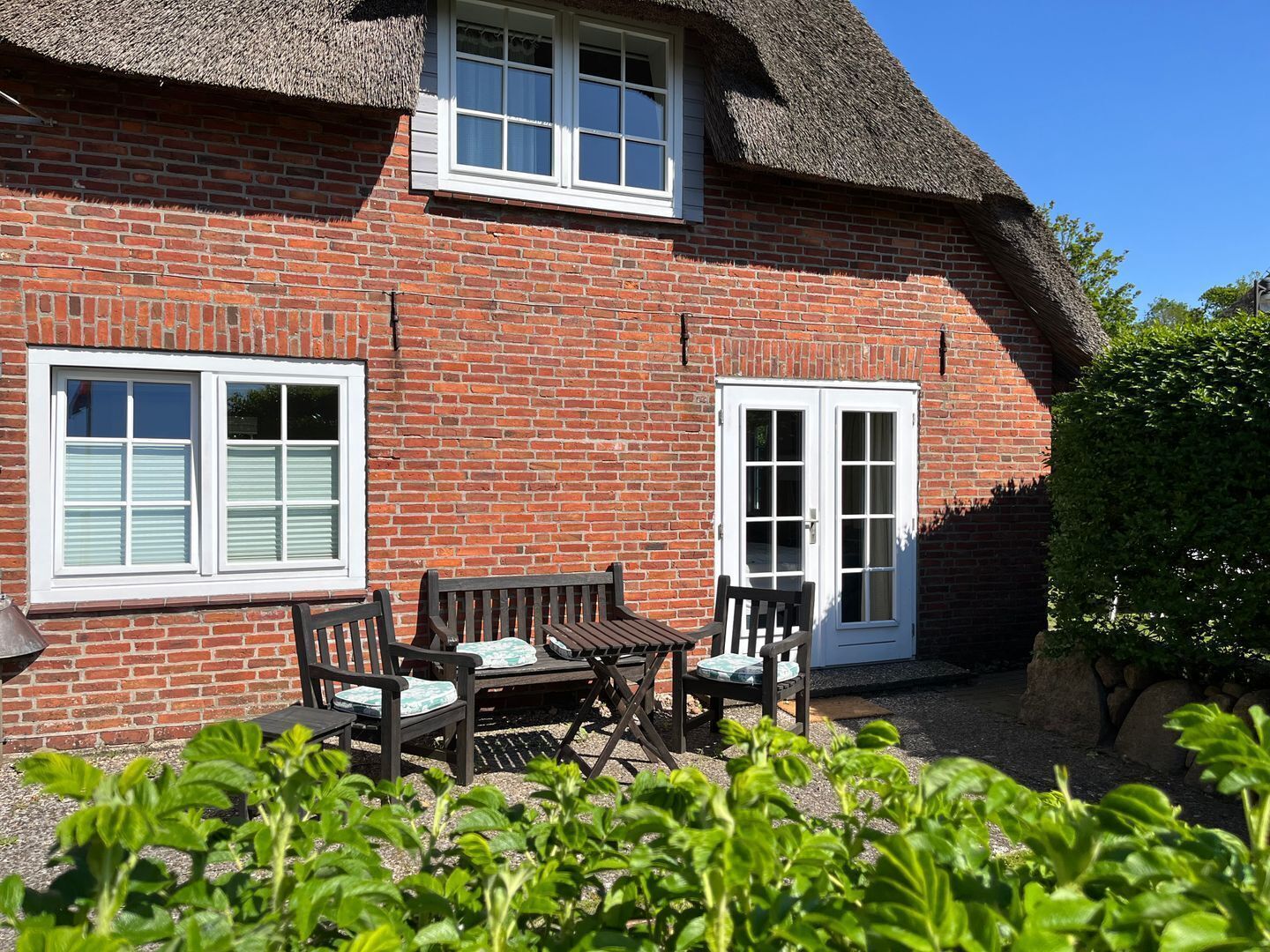 A brick house with a thatched roof, white-framed windows, and a door. There is a small patio with a table and chairs.