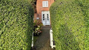 Une maison en briques rouges, avec une porte blanche et une petite fenêtre, entourée de haies soigneusement taillées.