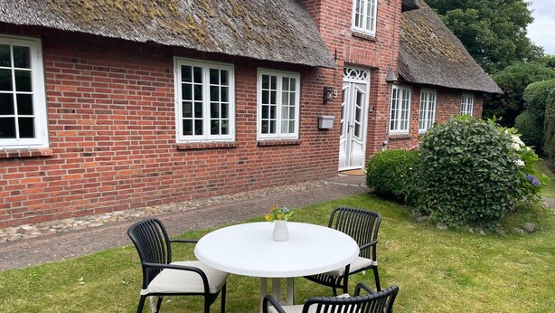 Une maison en briques avec un toit de chaume, une table ronde entourée de chaises et un petit jardin.