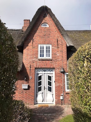 A red brick house with a thatched roof, a white double door, and a small window above the door.