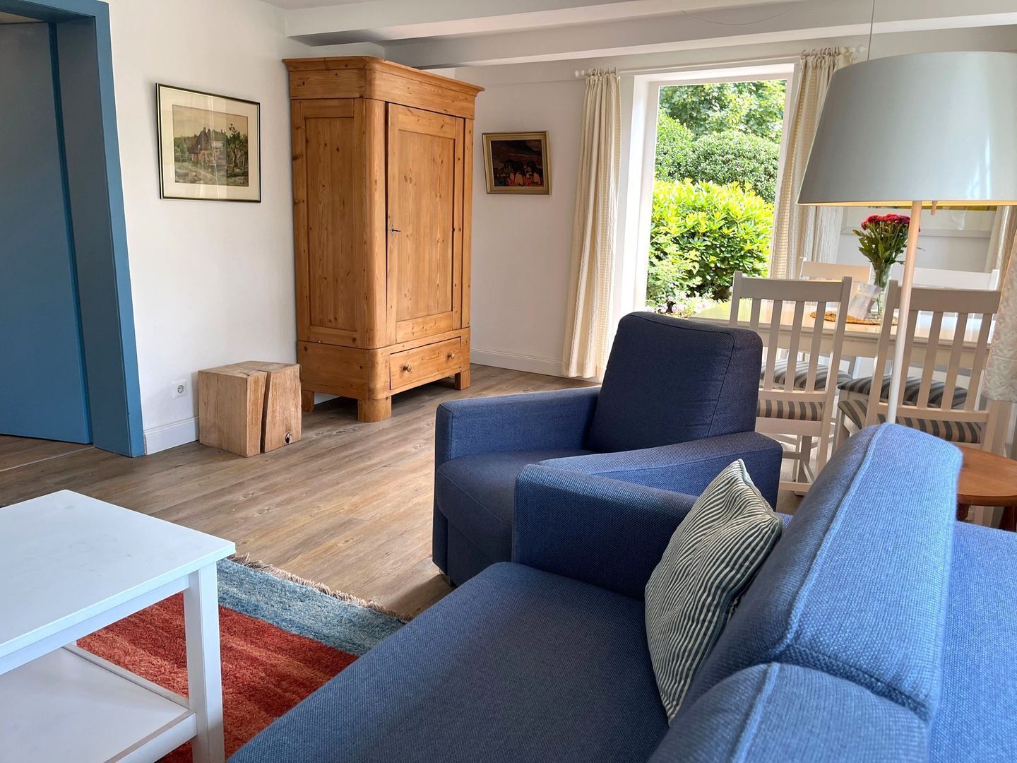 A living room with a blue sofa, a wooden cabinet, a white table, and a window with a view of greenery.