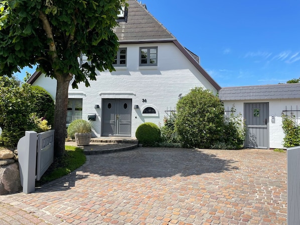 A white house with a grey door, number 36, and a small garden with a tree and bushes.