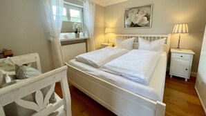 A bedroom with a double bed, two bedside tables, a window with curtains, a radiator, a wall-mounted painting, and a wooden chair. - First floor, 50sqm for up to 2 persons - Haus Thomsen (Sylt)