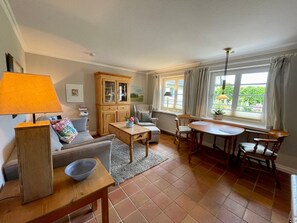 A cozy living room with a sofa, a wooden cabinet, a dining table with chairs, and a window with curtains. - First floor, 50 m² for up to 2 people - Thomsen House (Sylt)