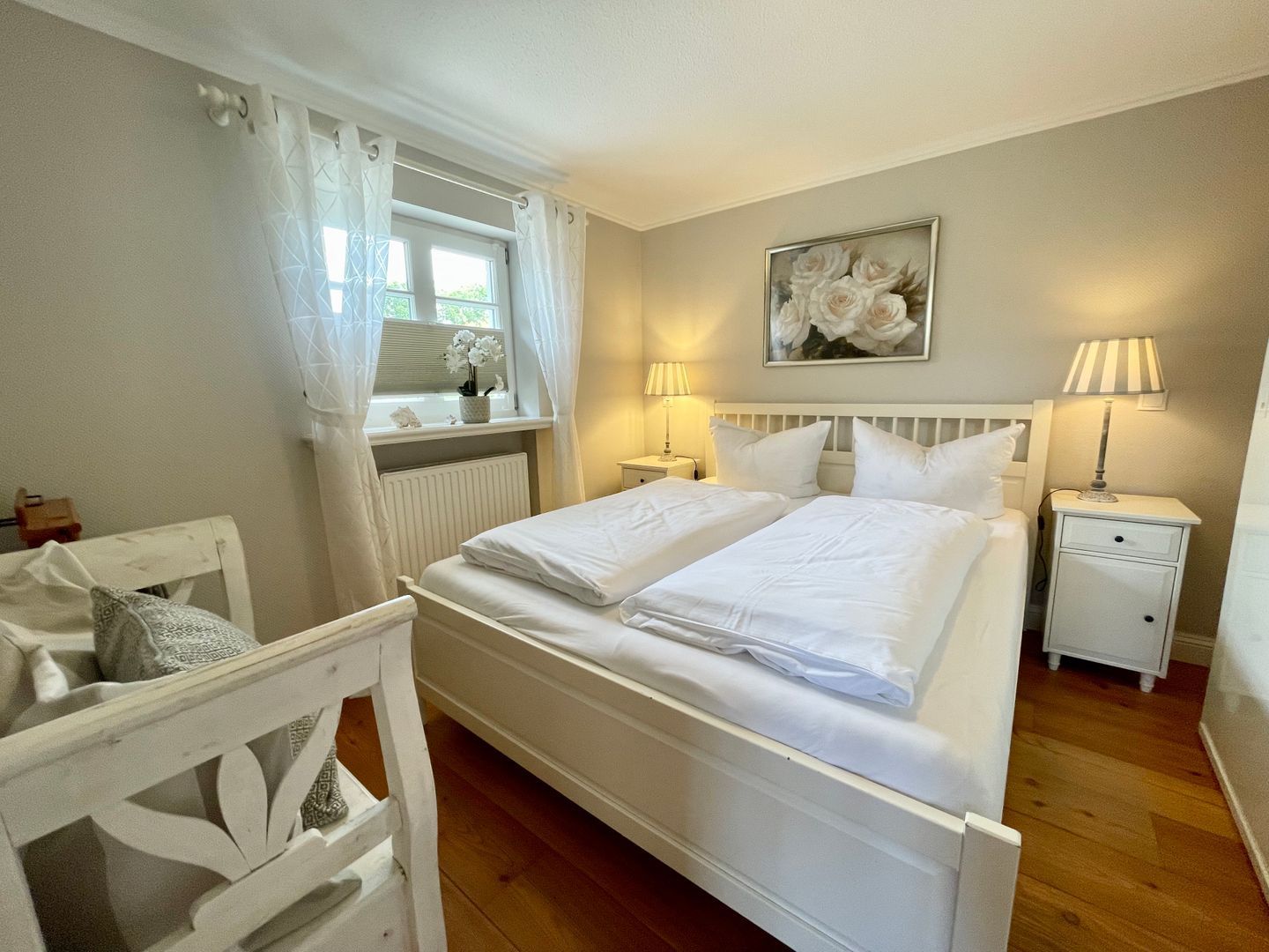 A bedroom with a double bed, two bedside tables, a window with curtains, a radiator, a wall-mounted painting, and a wooden chair.