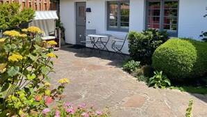 A stone patio with a table and chairs, surrounded by greenery and a white building with windows. - First floor, 50sqm for up to 2 persons - Haus Thomsen (Sylt)