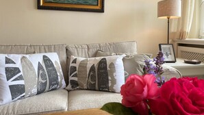 A living room with a beige sofa, patterned cushions, a framed picture of a ship, a lamp, and a vase with flowers. - Top floor, 50sqm for up to 2 persons - Haus Thomsen (Sylt)