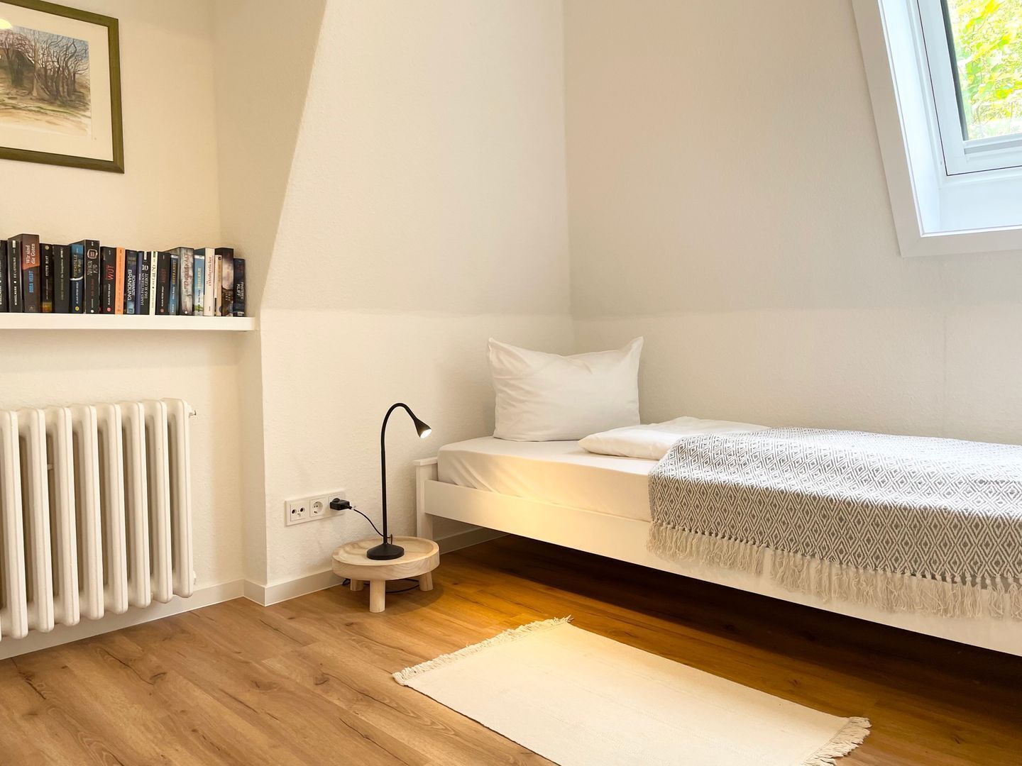 A small, well-lit bedroom with a single bed, a bedside table, a lamp, a radiator, and a bookshelf with books.