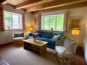 Ein gemütliches Wohnzimmer mit einem blauen Sofa, zwei Korbstühlen, einem Holztisch und zwei mit Vorhängen versehenen Fenstern.