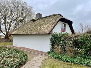 A small white cottage with a thatched roof and a red window, surrounded by greenery and a stone pathway.
