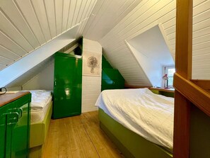 A dormitory room with a sloped ceiling, green wardrobe, wooden dresser, and a bed with white bedding.