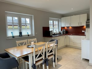 A modern kitchen with white cabinets, a wooden dining table, and two windows with blinds.