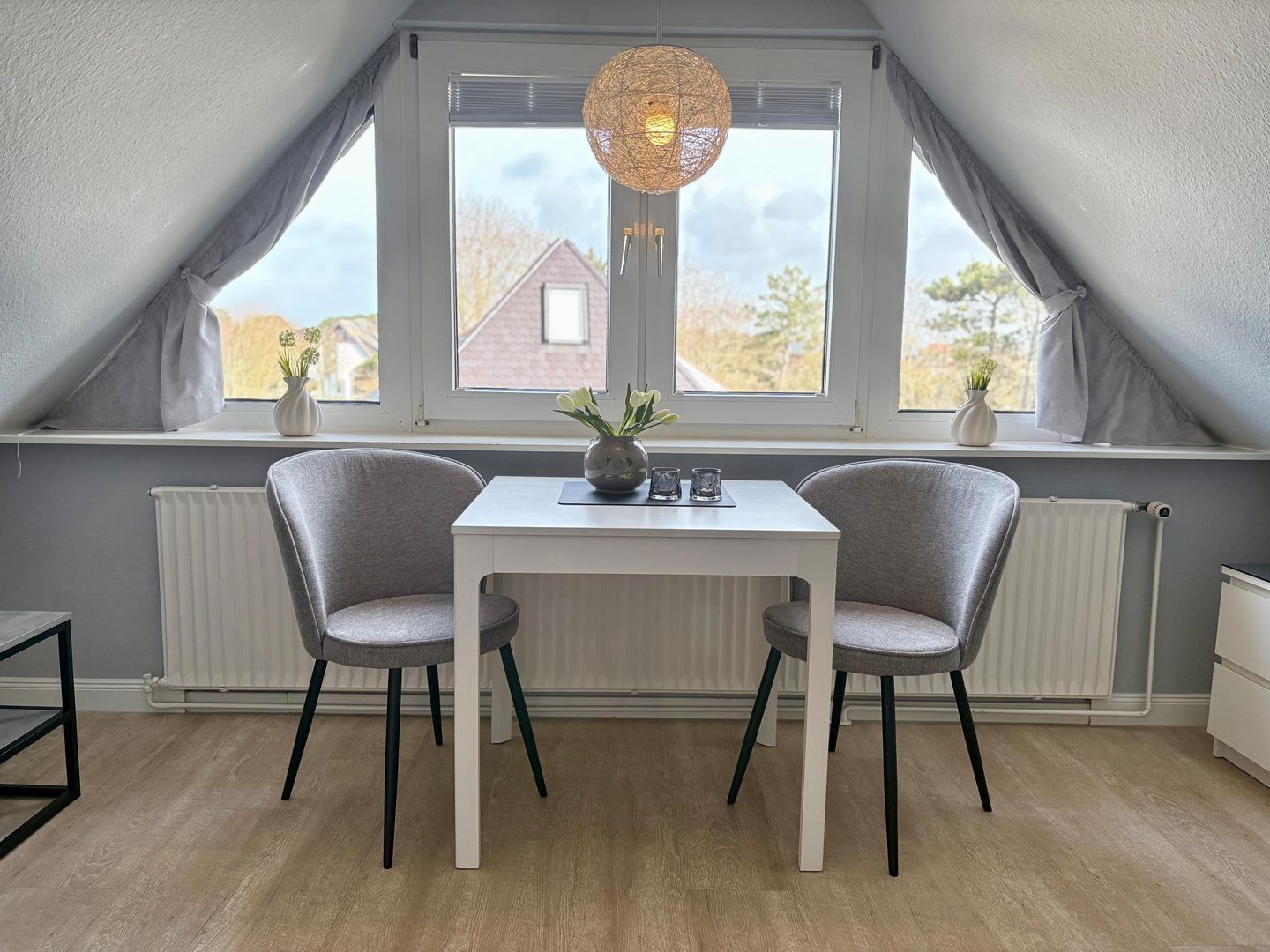 Une pièce comprenant une table et deux chaises, une fenêtre avec des rideaux, un radiateur et un luminaire suspendu.
