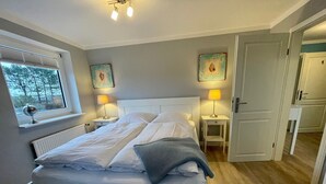 Een netjes opgemaakt bed in een kamer met een raam, twee nachtlampjes en een deur naar een andere ruimte.