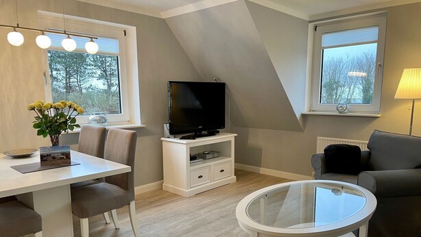 Een moderne woonkamer met een eethoek, een flatscreen televisie op een witte standaard, een salontafel met glazen blad, en een grijze bank.
