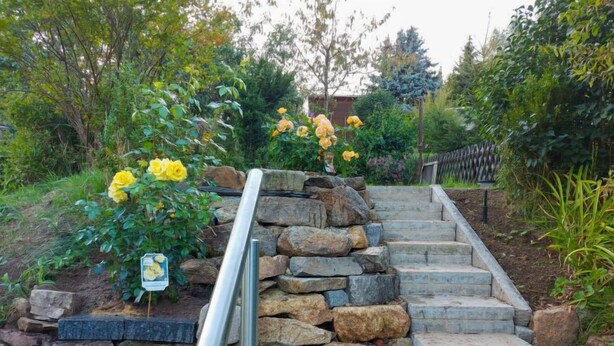Een stenen trap met een metalen leuning die leidt naar een tuin met gele bloemen en een bord.