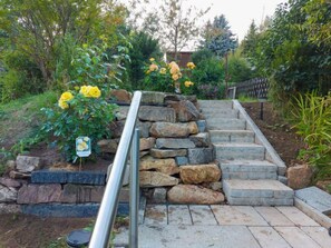 Un escalier en pierre avec une rampe métallique mène à un espace jardin agrémenté de fleurs jaunes et d’un panneau.