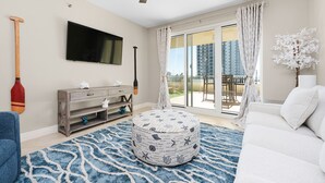 A living room with a flat-screen TV, a wooden TV stand, a round ottoman with nautical prints, a blue patterned rug, a ceiling fan, and a sliding glass door leading to a balcony. - Ariel Dunes I 303 * Last minute savings. Walk to Beach! Salt Water Vacations (Destin)