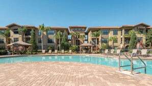 A pool area with a brick deck, lounge chairs, and a building with multiple balconies and palm trees. - 9 miles to Disney, washer dryer, full kitchen, private patio, 2 community pools (Davenport)