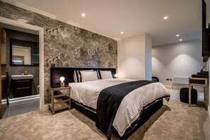 A modern bedroom with a large bed, a nightstand with a lamp, a mirror, and a dark headboard.