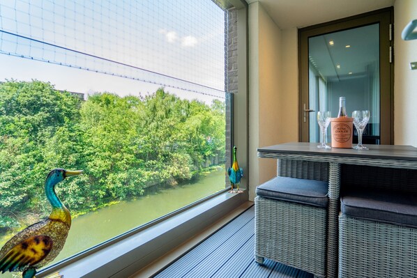 A room with a view of a river and trees, a wicker sofa, a table with a bottle and glasses, and a decorative bird.