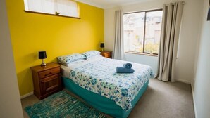 Een slaapkamer met een bed, nachtkastje, lamp, raam met gordijnen en uitzicht op gebouwen.