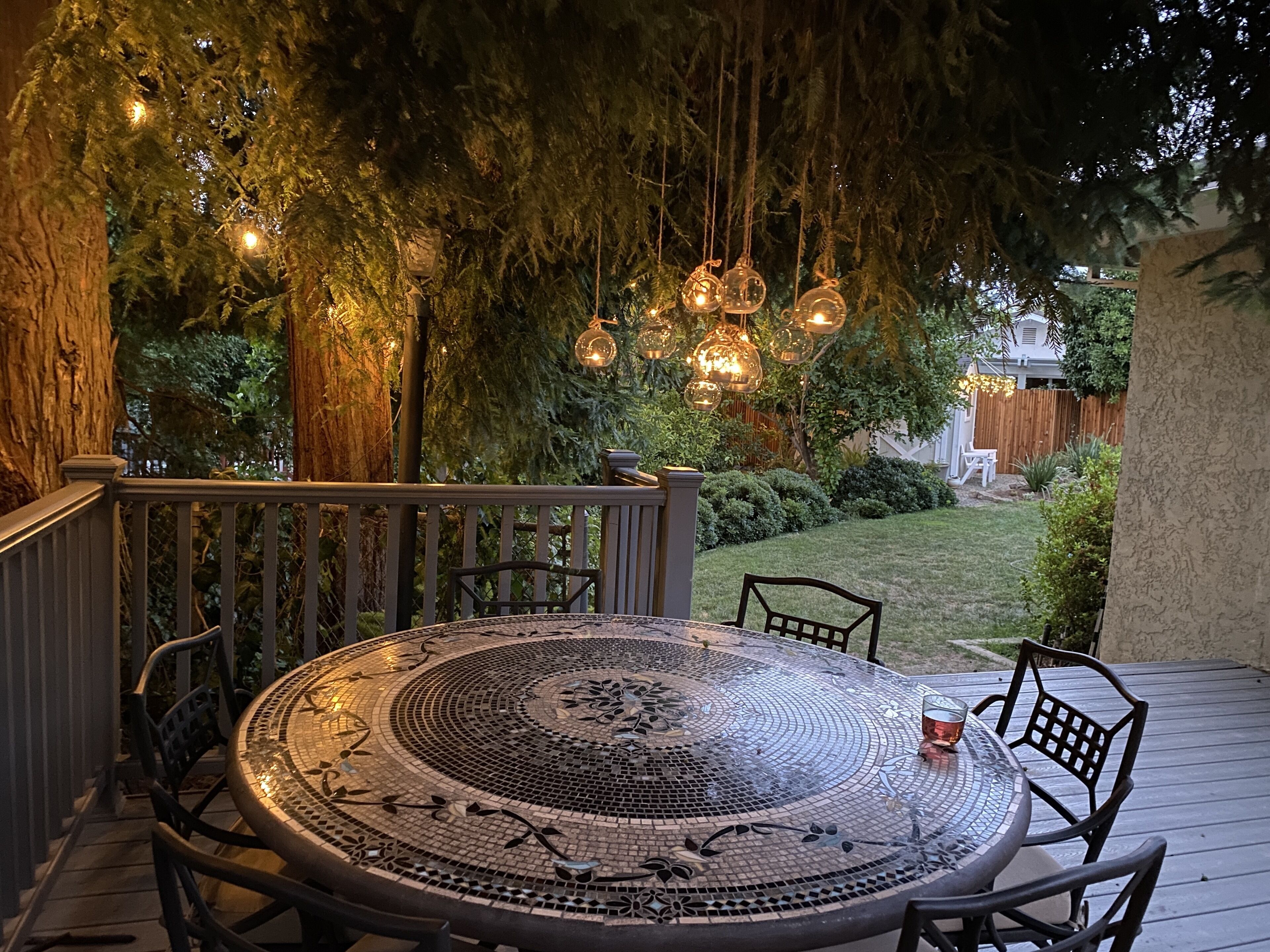 A round outdoor table with mosaic tile design, surrounded by chairs, under a tree with hanging lights.