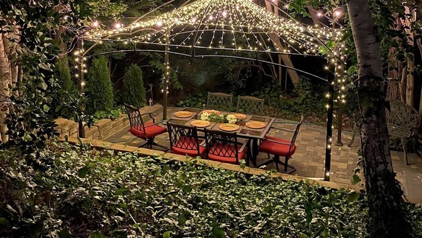 Een buitenruimte met een overdekte eettafel en stoelen, omgeven door groen en sfeerverlichting.
