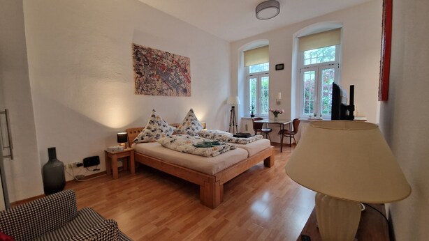 Ruime slaapkamer met een houten bed, een bureau en een stoel. Grote ramen zorgen voor natuurlijk licht en uitzicht op groen buiten.