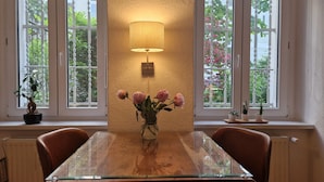 Een eethoek met een glazen tafel, stoelen, een vaas met bloemen, een wandlamp en ramen met uitzicht op groen.
